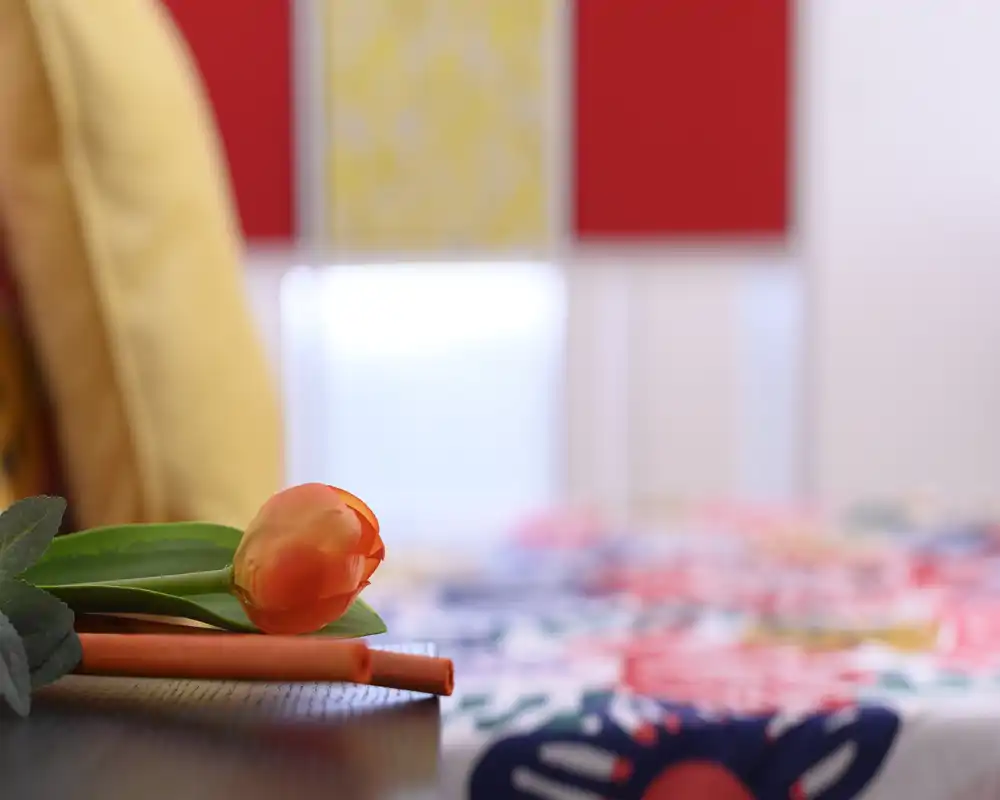 Toma en primer plano con estilo de vida en un dormitorio que muestra un tulipán decorativo sobre una superficie de mesa/mesita con una funda colorida desenfocada al fondo. La imagen enfatiza el detalle y la atmósfera más que la cama completa.