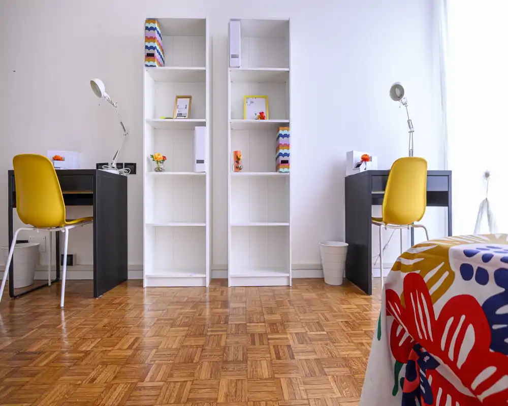 Rincón de dormitorio moderno y luminoso con suelo de parquet, dos escritorios con sillas amarillas y estanterías blancas abiertas; se ve parcialmente una colcha colorida a la derecha.