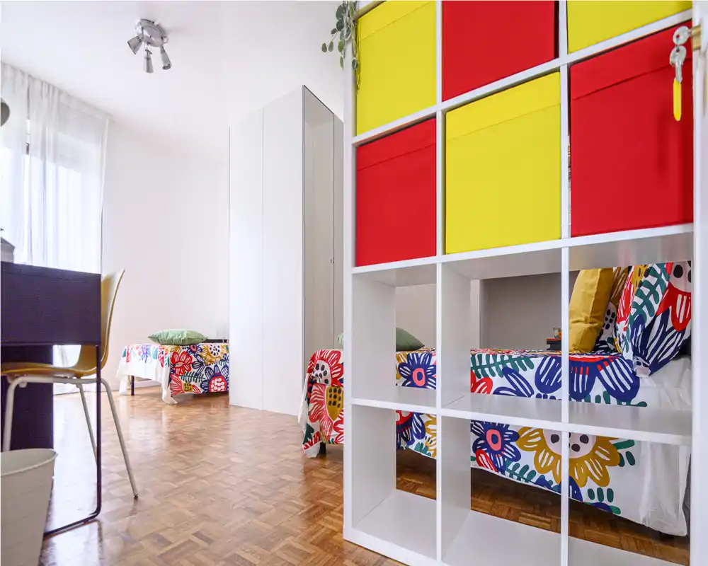 Dormitorio luminoso y colorido con dos camas individuales separadas por estanterías; luz natural abundante y ropa de cama vibrante que crea un ambiente alegre.