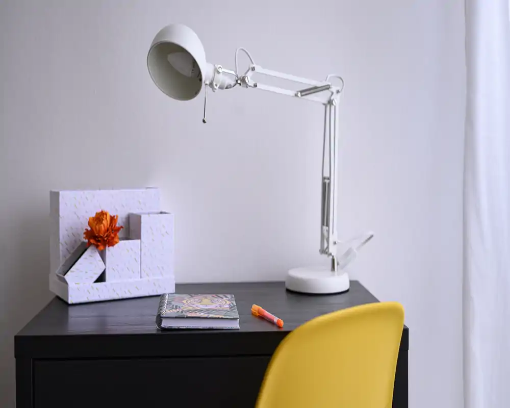 Rincón de trabajo ordenado con una lámpara de escritorio blanca moderna, caja decorativa, cuaderno y una silla amarilla brillante — ideal para un espacio de trabajo compacto.