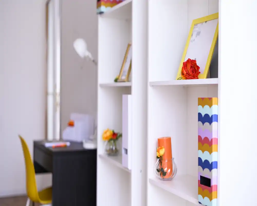 Zona de estar luminosa y moderna con estanterías blancas y decoración colorida; se aprecia un pequeño escritorio y silla, ideal como rincón de trabajo.