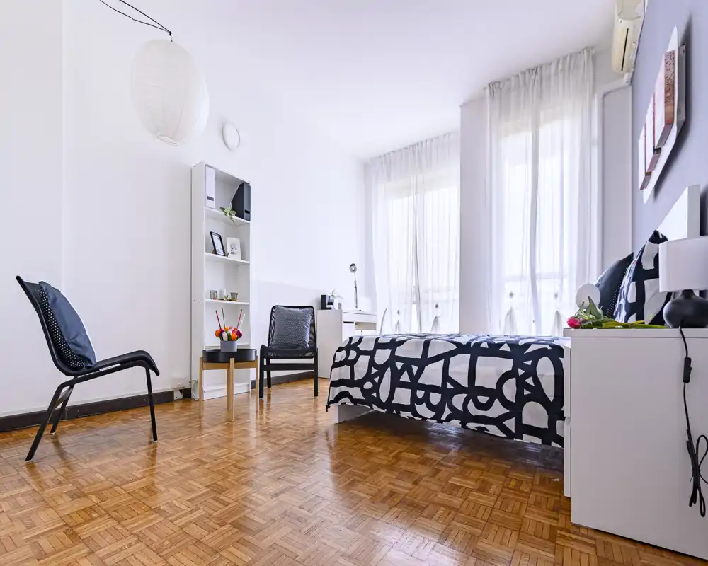 Camera da letto moderna e luminosa con copriletto a motivo, grandi finestre con tende leggere e mobili ordinati: perfetta per annunci che valorizzano spazio e luce.