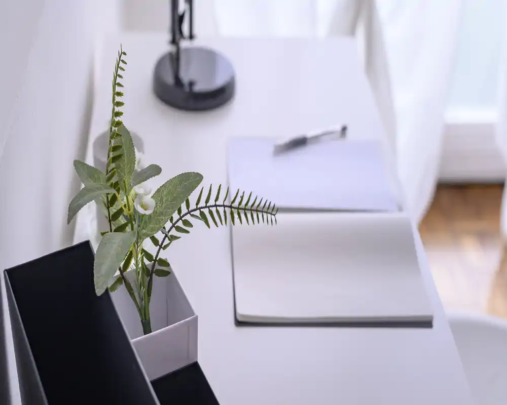 Angolo scrivania luminoso e minimal con taccuino, penna e piccola pianta vicino a una lampada — un'area di lavoro tranquilla con luce naturale.