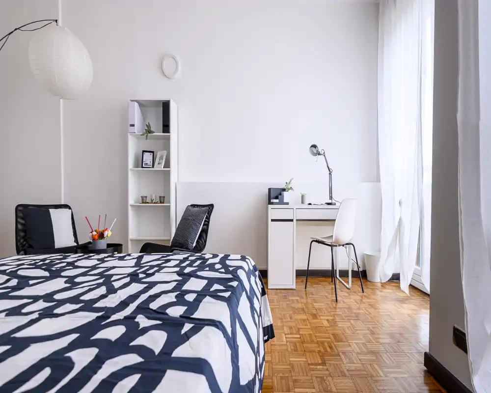 Camera da letto luminosa e moderna con letto matrimoniale dal copriletto a motivo, scrivania vicino alla finestra e arredi essenziali e di design, perfetta per un annuncio.