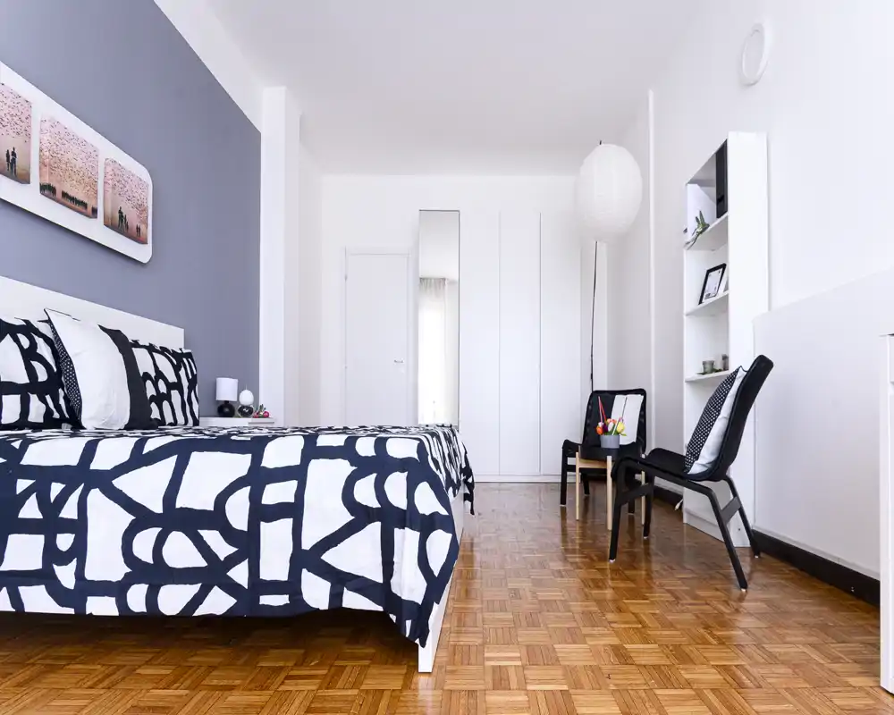 Camera da letto luminosa e moderna con un ampio letto a fantasia, pavimento in parquet ordinato e arredi minimalisti che creano un'atmosfera elegante e rilassante.