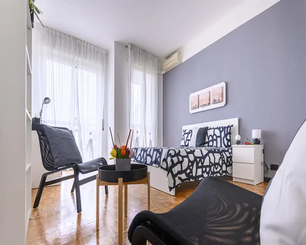 Camera da letto moderna e luminosa con biancheria a motivo, ampie finestre con tende leggere e comode sedute — perfetta per un annuncio di affitto elegante.
