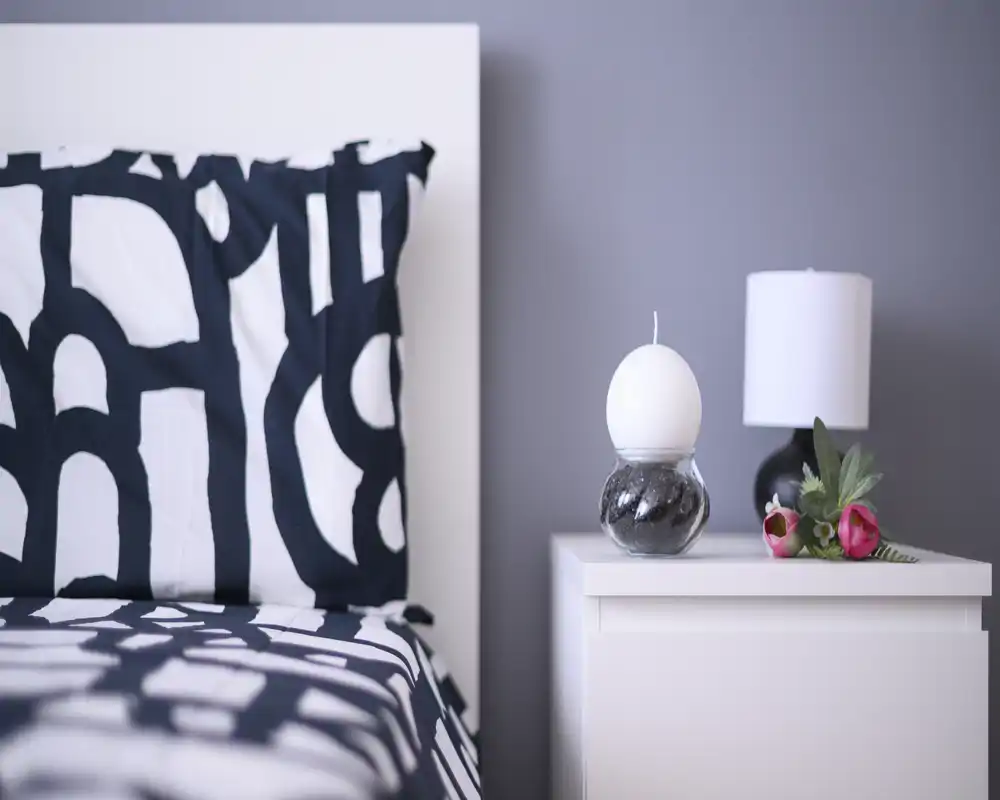 Camera ravvicinata di una camera da letto luminosa: cuscino a motivo e parte del letto accanto a un comodino bianco con lampada e candela decorativa — moderno e minimale.