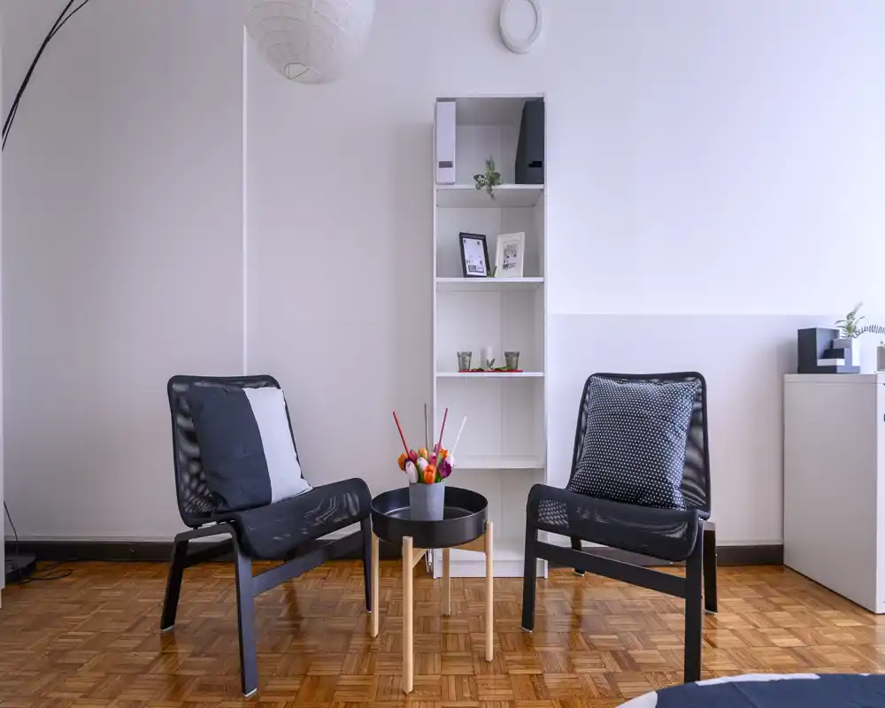 Zona giorno luminosa e minimal con due poltrone moderne, un tavolino rotondo con fiori e una libreria alta contro una parete chiara.