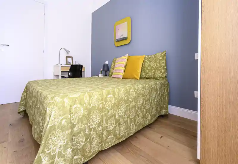 Well-lit, styled bedroom with a green patterned bedspread, decorative cushions and a small desk in the corner. The image highlights the bed and cozy decor, suitable for rental listing photos.