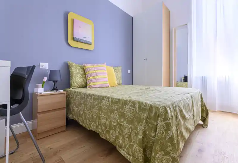 Bright, well-styled bedroom with a double bed dressed in patterned olive-green linens, colorful cushions, bedside table with lamp and a wardrobe near a sheer-window allowing abundant natural light.