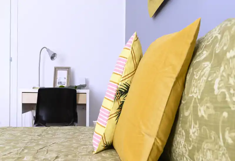 Bright, styled bedroom close-up showing decorative pillows and part of the bed with a small desk in the background — cozy and well-lit space, ideal for listings highlighting comfort and design.