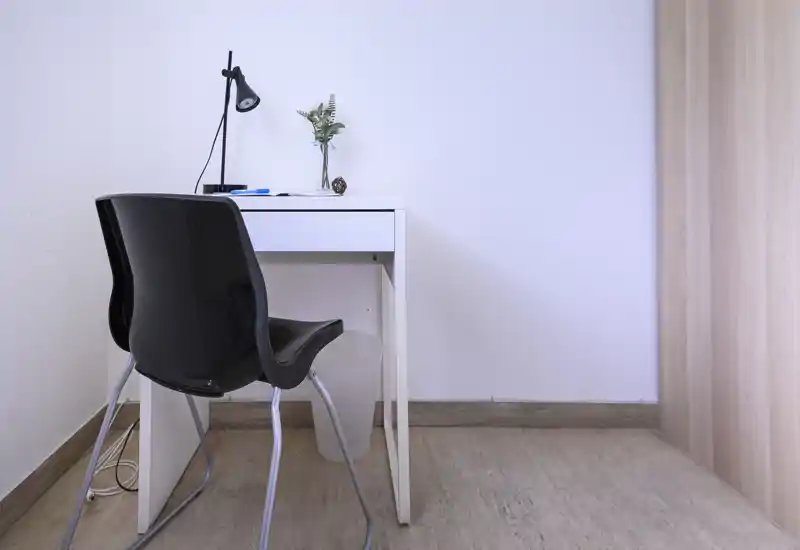 Espacio de trabajo compacto con una mesa blanca, silla negra y lámpara de escritorio; decoración mínima con un pequeño jarrón. Rincón luminoso, limpio y funcional ideal para teletrabajo o estudio.