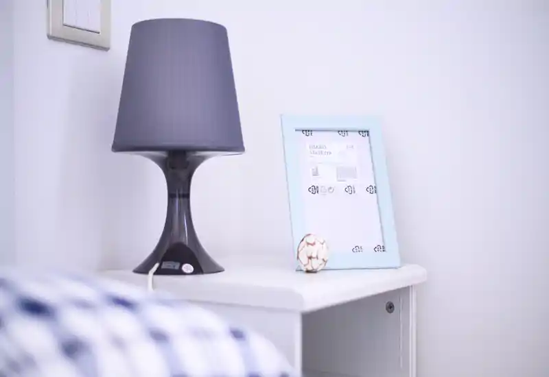 Primo piano di un comodino con lampada moderna, cornice decorativa e piccolo oggetto; una porzione del letto è visibile in basso a sinistra, indicando un dettaglio della camera da letto.