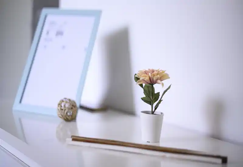 Superficie luminosa e minimal vicino al letto o un comò con un piccolo fiore in vaso e oggetti decorativi, che suggerisce un angolo tranquillo della camera da letto.