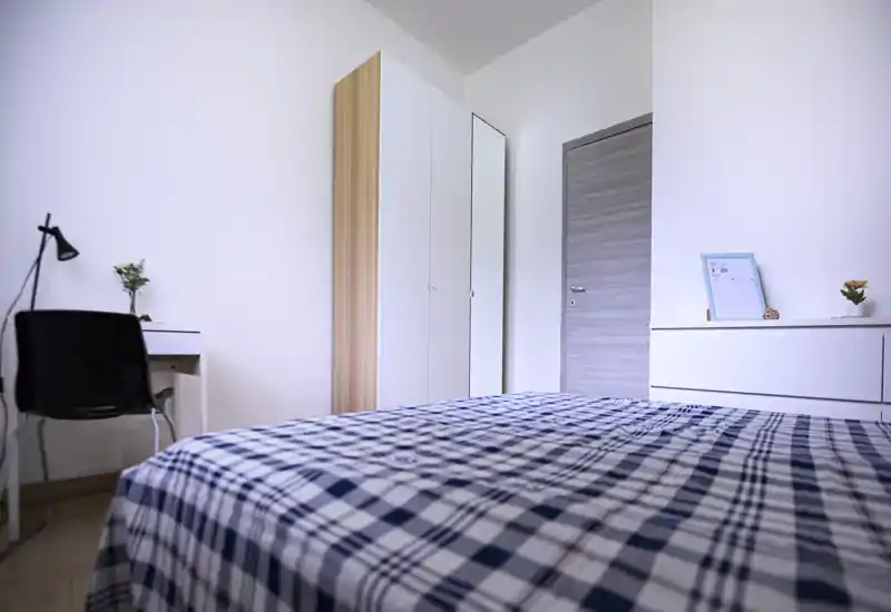 Camera da letto luminosa e minimal con copriletto a quadri, armadio con specchio e una piccola scrivania — ben illuminata, ordinata e arredata in modo semplice.
