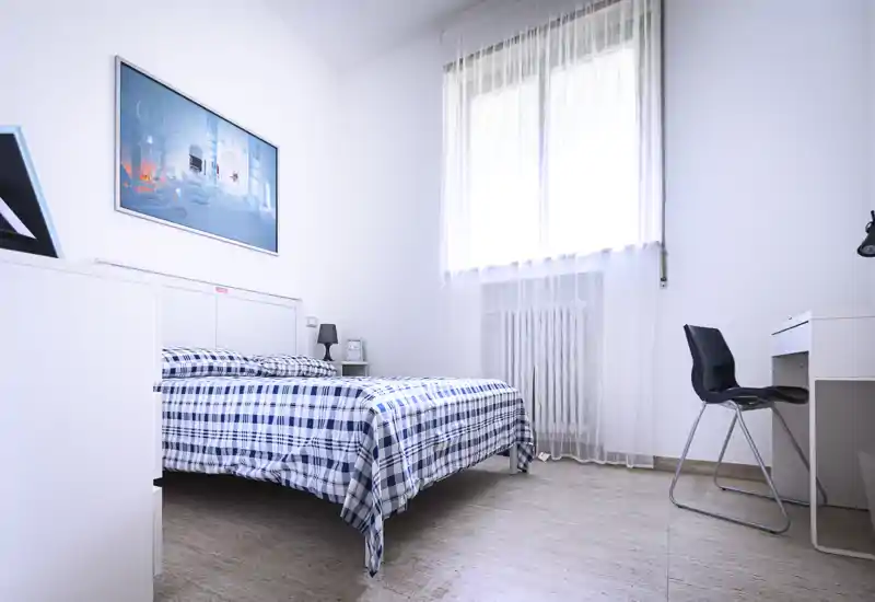 Camera da letto luminosa e minimale con letto matrimoniale con copriletto a quadri blu, comodino con lampada e scrivania con sedia vicino alla finestra. La luce naturale diffusa e l’arredamento essenziale creano un’atmosfera tranquilla e ariosa, perfetta per annunci.