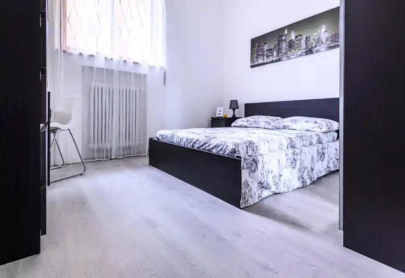 Bright, modern bedroom with a dark wooden bed, patterned bedding, bedside table and cityscape artwork — clean, well-lit and ready for guests.