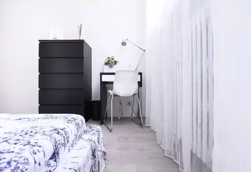 Minimalist bedroom corner with a patterned bed in the foreground, a tall dark chest of drawers, and a compact desk with lamp by a sheer-curtained window.
