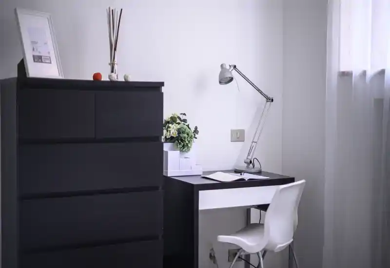 Minimalist study corner with a compact desk, task lamp and chair beside a tall storage chest; bright natural light and simple decorative touches create a calm workspace.