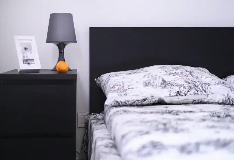 Bright, modern bedroom close-up featuring a neatly made bed with patterned linens and a dark headboard; a minimal nightstand with lamp and small decor completes the scene.