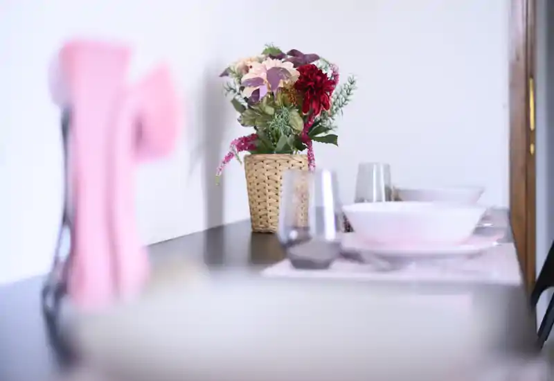 A softly lit dining setup with a woven floral centerpiece and place settings on a narrow table, shown with intentional shallow depth of field for a cozy, styled look.