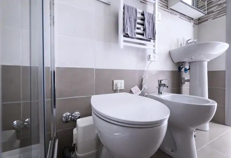 Clean, modern bathroom with a toilet, bidet, pedestal sink and towel radiator; neutral beige tiles and a glass shower door create a bright, fresh look.