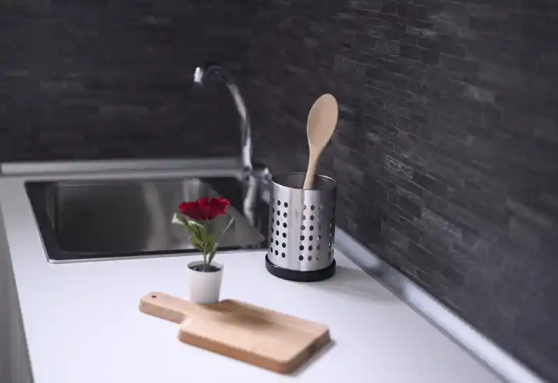 Minimal modern kitchen counter with a stainless sink, utensil holder with a wooden spoon, small cutting board and a potted decorative flower on a clean white surface.