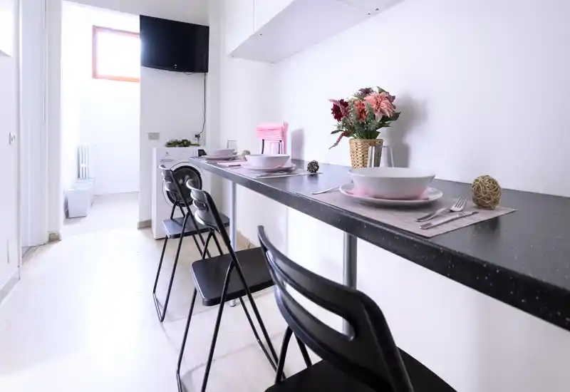 Bright, modern kitchenette with a long breakfast bar set for dining, black stools and decorative flowers—ideal for quick meals and social breakfasts.