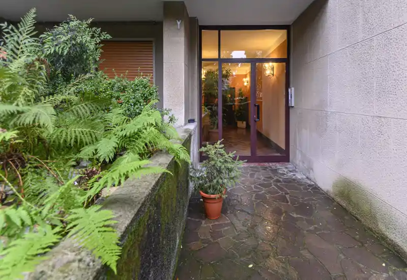 Exterior entrance path with potted plant and glass door leading into a well-lit lobby; stone paving and planted greenery create a welcoming approach.