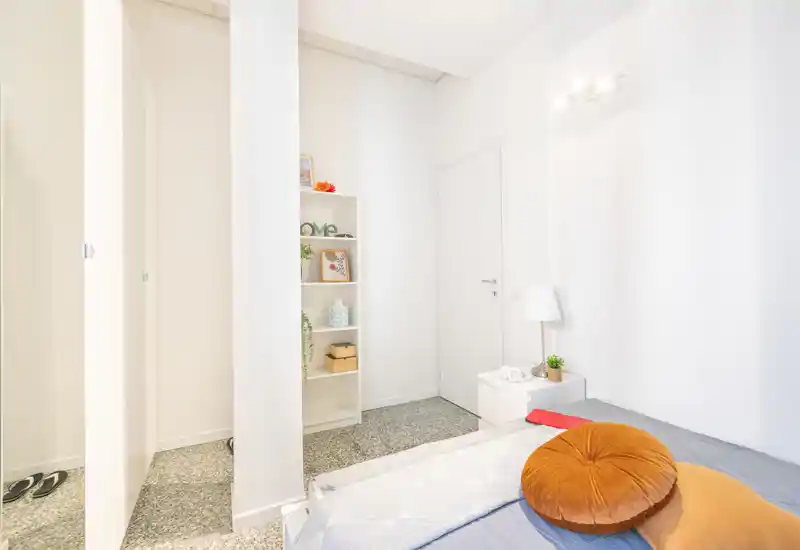 Camera da letto luminosa e minimal con letto ben rifatto, cuscini tondi decorativi e un piccolo comodino; pareti bianche e scaffale che donano un senso di freschezza.