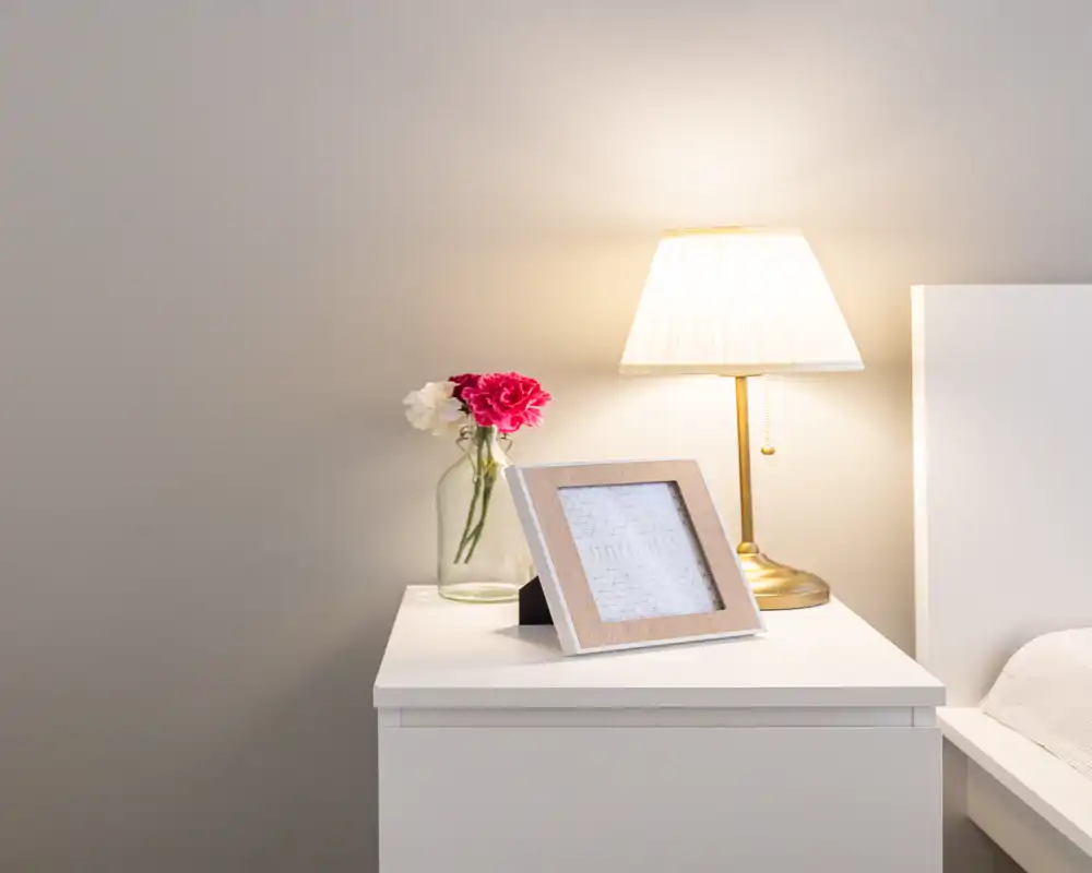 Angolo di camera luminoso e minimal con comodino bianco, lampada, fiori freschi e cornice; una piccola porzione del letto/testata è visibile sul bordo destro.