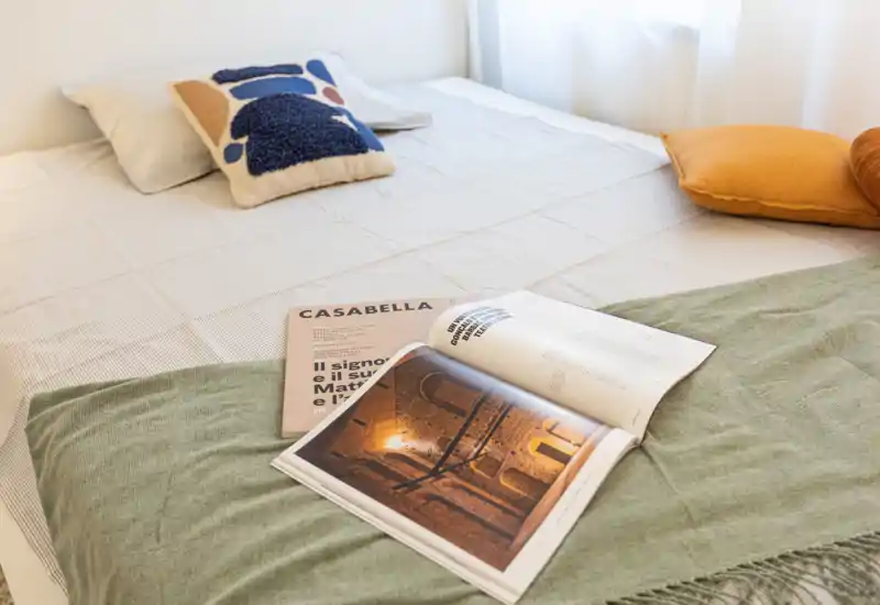 Camera da letto luminosa e ben arredata con un letto rifatto, cuscini decorativi e una rivista aperta sul copriletto, che trasmette un’atmosfera accogliente e moderna.