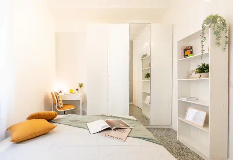 Camera da letto luminosa e moderna con letto in ordine, cuscini color senape, una scrivania compatta e un armadio con anta a specchio — ariosa e ben arredata, ideale per annunci.