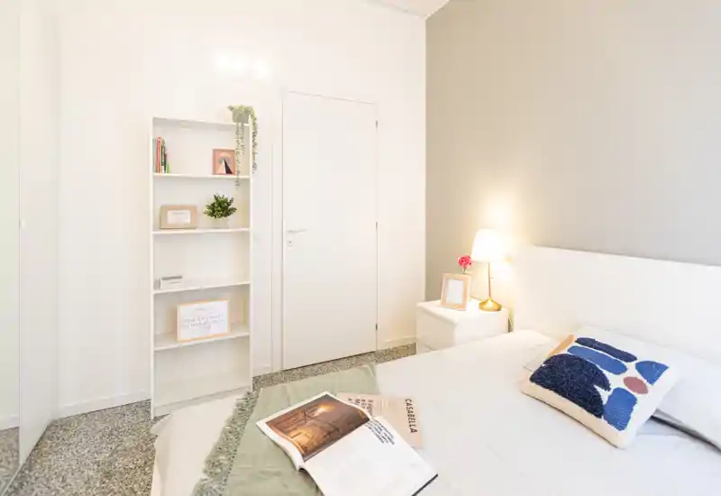 Camera da letto luminosa e moderna con letto ben rifatto, cuscini decorativi e comodino con lampada, arredata per un aspetto accogliente.