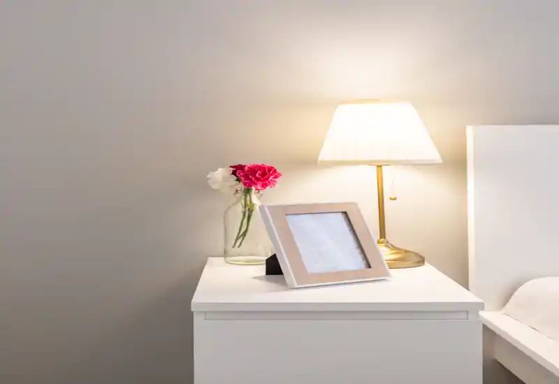 Angolo di camera luminoso e minimal con comodino bianco, lampada, fiori freschi e cornice; una piccola porzione del letto/testata è visibile sul bordo destro.