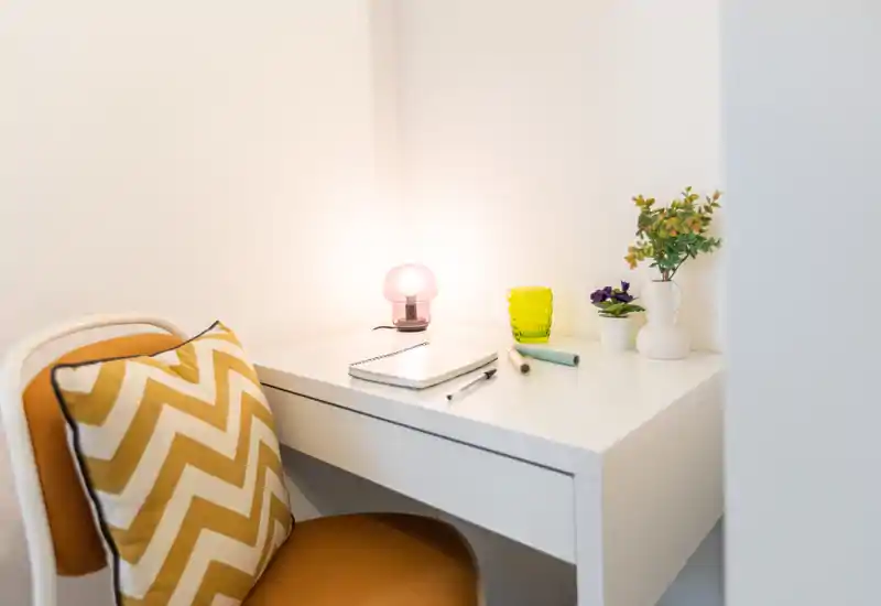 Angolo studio luminoso e curato con scrivania bianca, lampada calda e cuscino decorativo che creano un ambiente accogliente.
