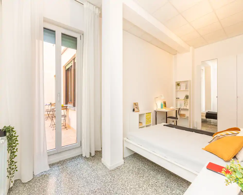 Camera da letto luminosa e moderna con letto matrimoniale, cuscini accoglienti e una scrivania. Ampie porte finestra si aprono su una piccola terrazza, garantendo molta luce naturale.
