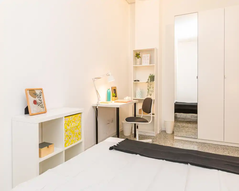 Camera da letto luminosa e moderna con letto ben rifatto in primo piano, una piccola scrivania con scaffale per lavoro o riporre oggetti e un armadio con specchio a tutta altezza.