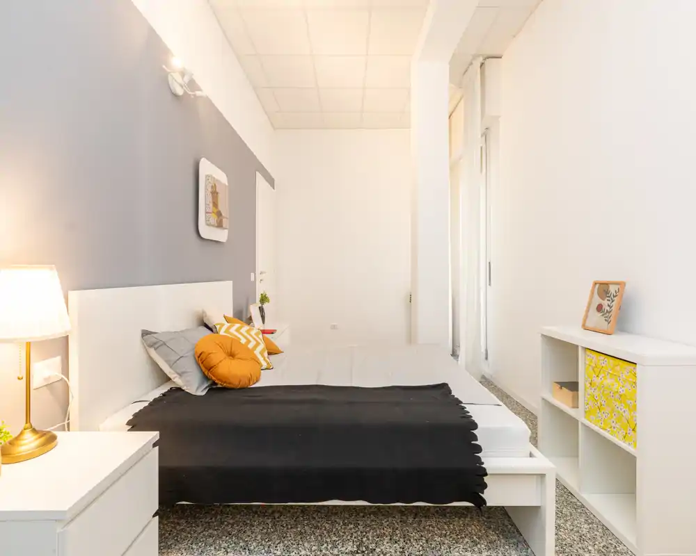 Camera da letto luminosa e moderna con ampio letto, cuscini decorativi e lampada da comodino. Palette neutra con dettagli caldi e mobili contenitori semplici, ideale per annunci.