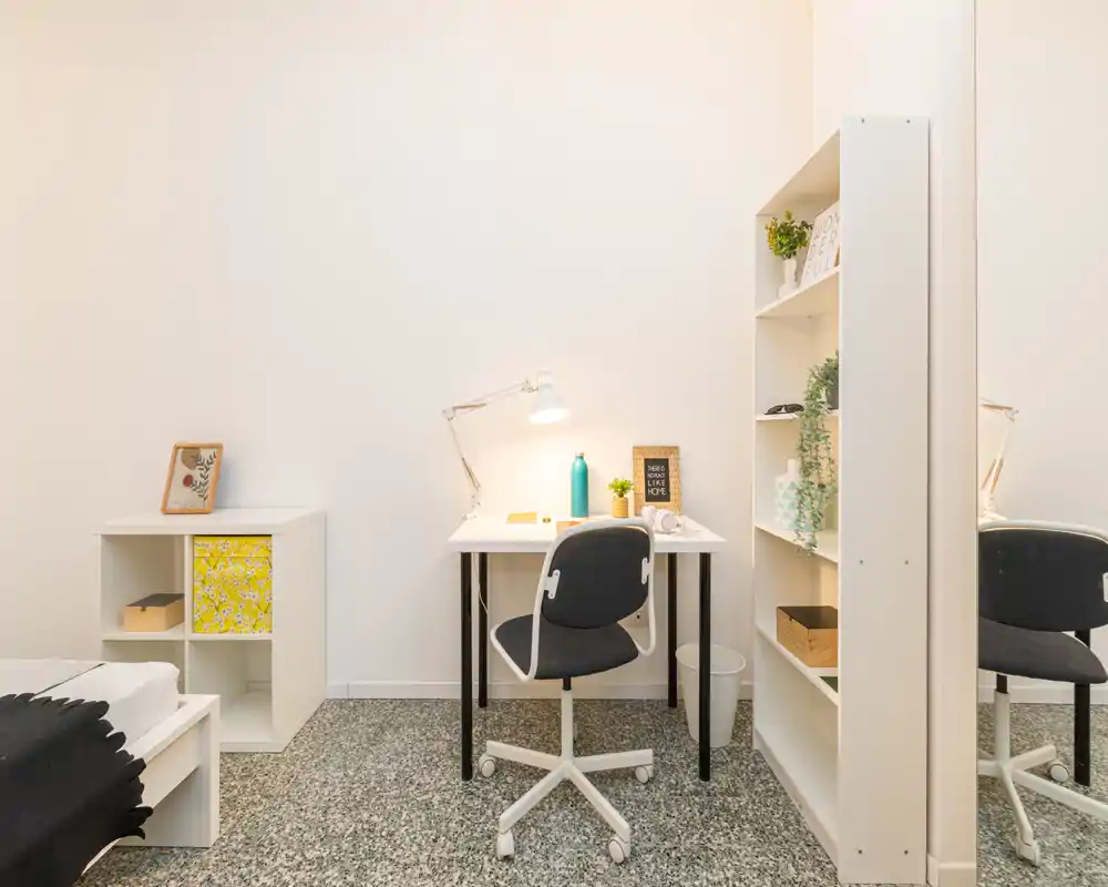 Angolo camera da letto luminoso e minimal con scrivania per lo studio, sedia ergonomica e scaffale. L'immagine mette in evidenza un'area di lavoro ordinata accanto al letto parzialmente visibile e al mobiletto.