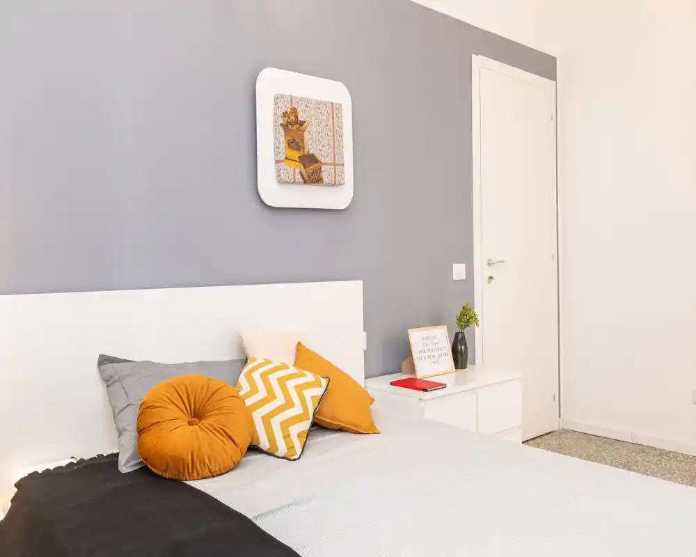 Camera da letto moderna e luminosa con testiera bianca, cuscini color senape e a zigzag e un comodino ordinato — perfetta per annunci accoglienti.