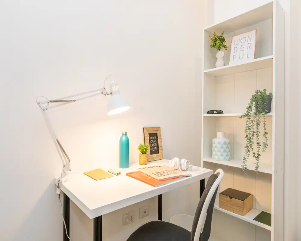 Angolo studio luminoso e ordinato con scrivania bianca, lampada regolabile, piante decorative e scaffalatura — ideale per lavorare o studiare da casa.