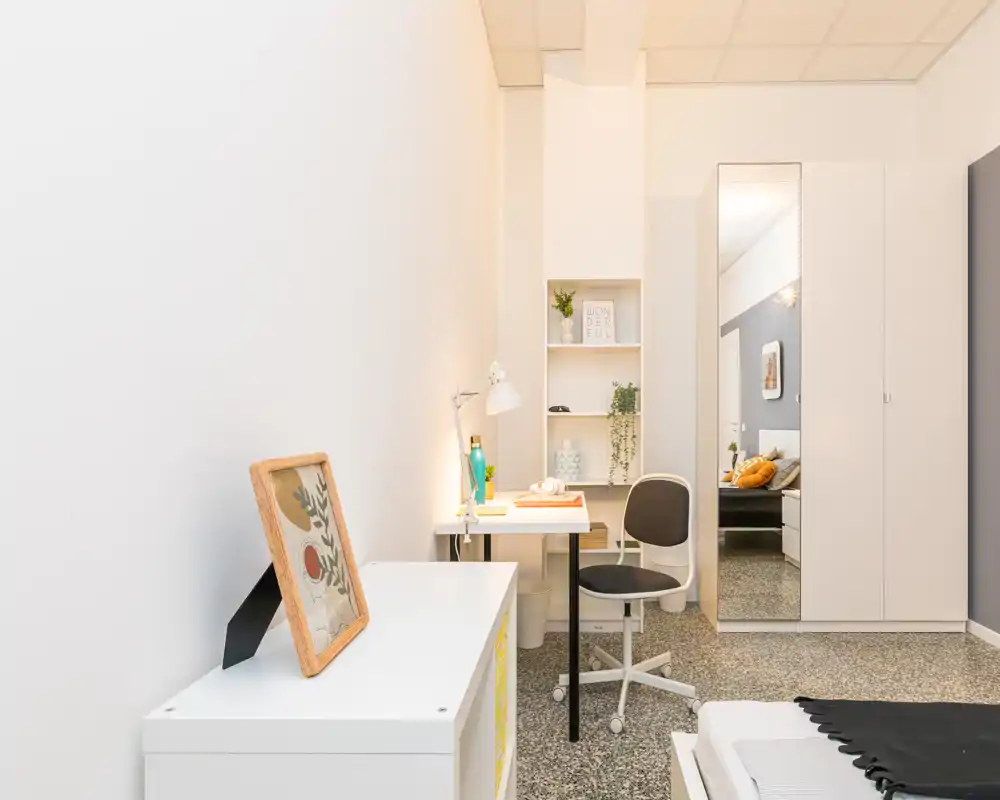 Angolo letto luminoso e minimal con scrivania e scaffalatura. L’immagine mostra una piccola zona studio, un armadio con specchio a tutta altezza e il bordo del letto, perfetta per annunci per una singola occupazione.