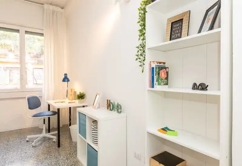 Angolo camera luminoso e minimal con una scrivania e scaffalatura — piccolo spazio lavoro ordinato vicino alla finestra con tende bianche e dettagli decorativi.