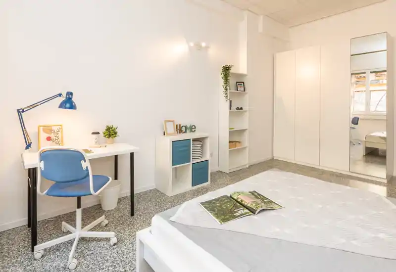 Camera da letto luminosa e ben arredata con un letto comodo in primo piano, una scrivania ordinata e mobili contenitivi, e un armadio con specchio che riflette luce.
