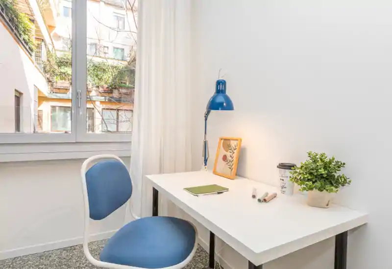 Angolo scrivania luminoso e minimal vicino a una grande finestra con sedia blu, lampada da tavolo, piccola pianta e stampa incorniciata — ideale per lavorare o studiare con luce naturale.