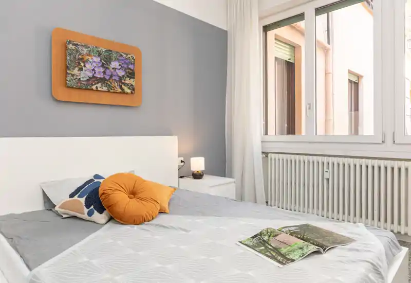 Camera da letto luminosa e curata con testiera bianca, cuscini decorativi e luce naturale da una grande finestra — accogliente e ben illuminata per annunci.