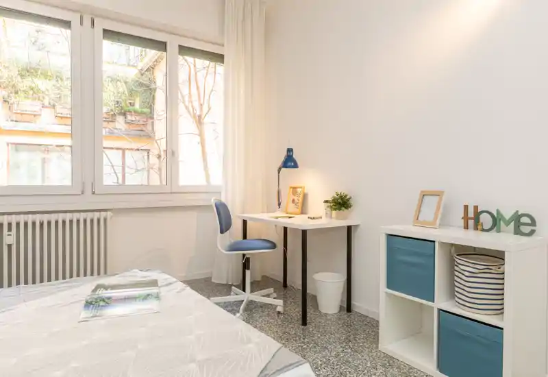 Camera da letto luminosa e minimale con grande finestra, scrivania semplice e dettagli accoglienti — ideale per una persona o per studenti.