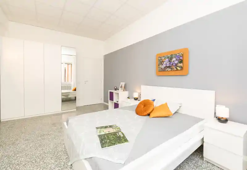 Camera da letto luminosa e moderna con struttura del letto bianca, cuscini arancioni decorativi e comodini minimal; parete grigia e armadio con anta a specchio donano un aspetto pulito e spazioso.