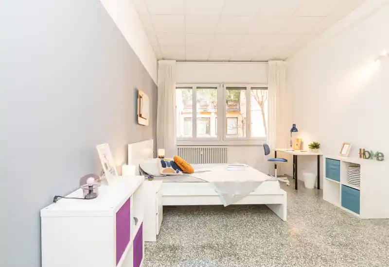 Camera da letto luminosa e ben arredata con letto matrimoniale centrale, cuscini decorativi, angolo studio vicino alla finestra e mobili contenitori moderni — perfetta per un affitto accogliente.